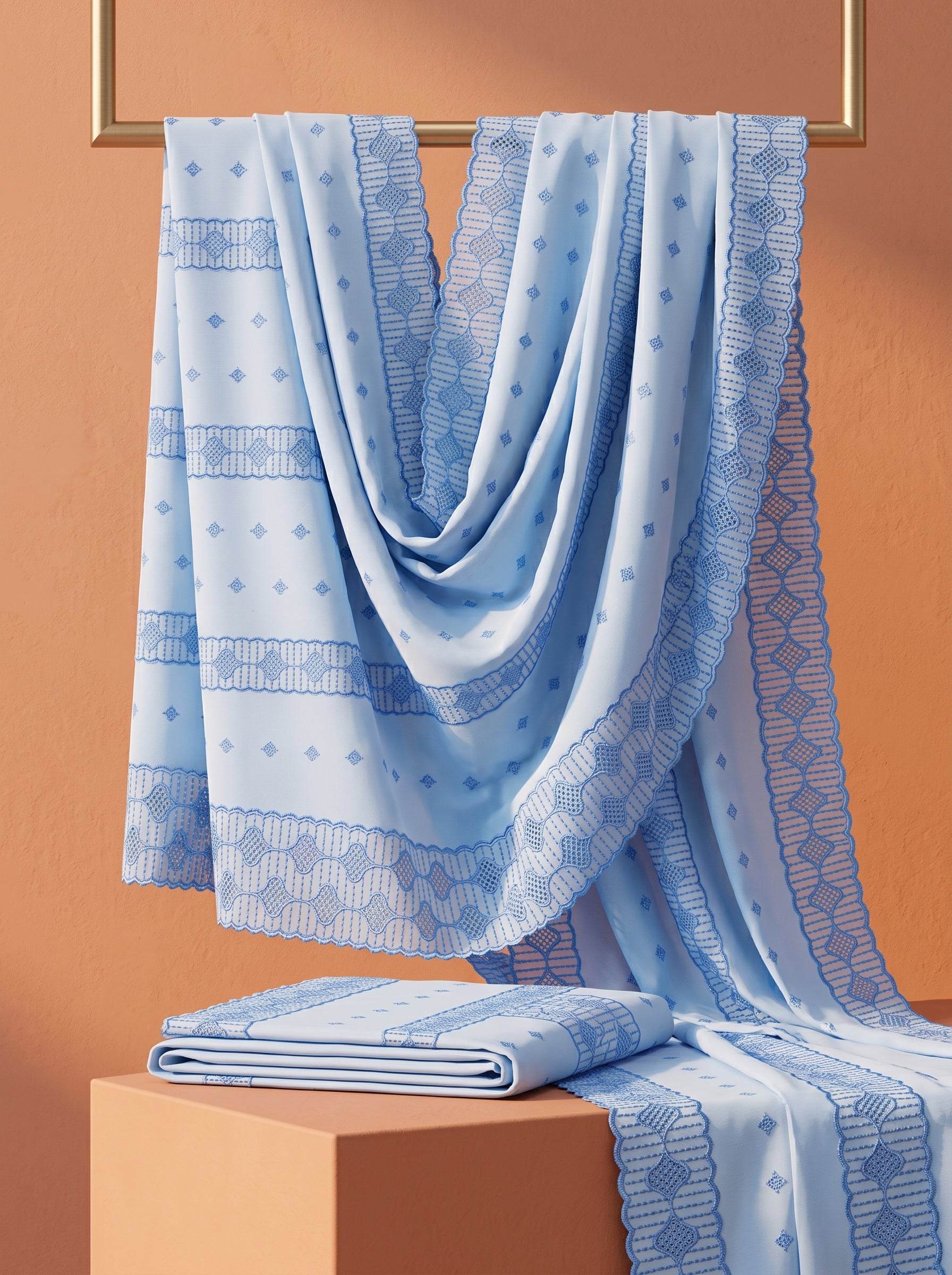 A light blue patterned fabric, likely a dupatta or scarf, draped elegantly over a gold-framed wall fixture, with a matching folded piece resting on a terracotta-colored pedestal against a warm terracotta background.